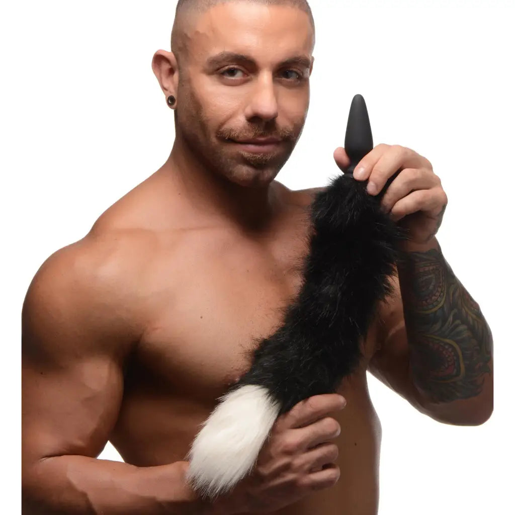 Muscular tattooed man holding a small anal plug with a fluffy black and white fox tail